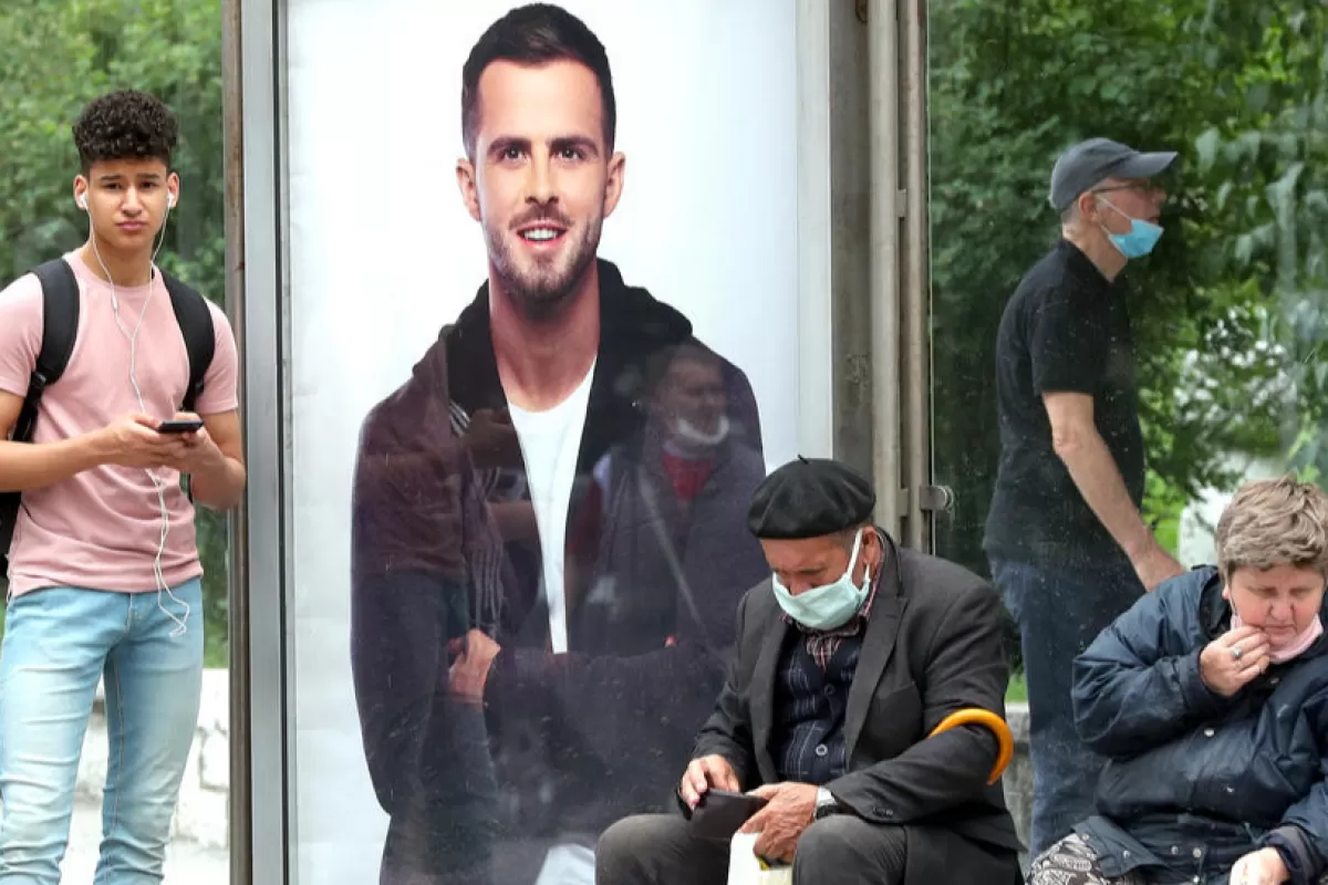 Oamenii care poartă măști faciale așteaptă în stația de tramvai, în fața unei imagini cu celebrul fotbalist bosniac Miralem Pjanic, în Saraievo, Bosnia și Herțegovina, 15 iunie 2020.